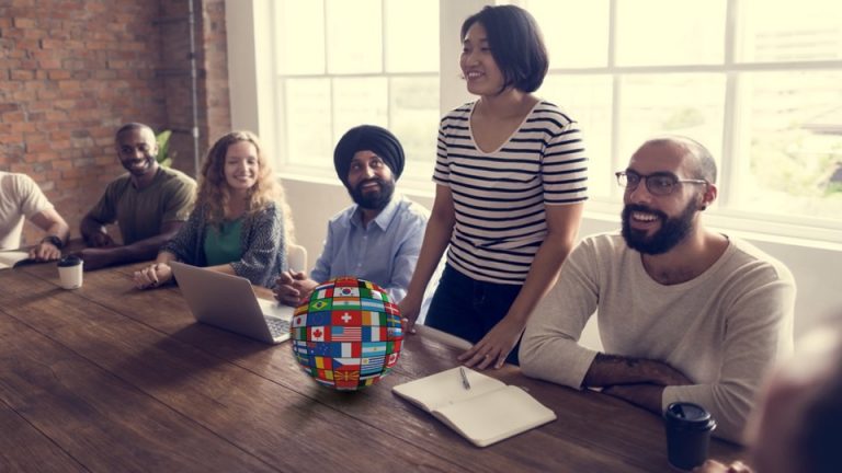 A woman stands at a team meeting. While others have laptops, coffee and notebooks, she has a globe