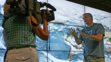 A man is interviewed by a cameraperson in front of a map of the world from space